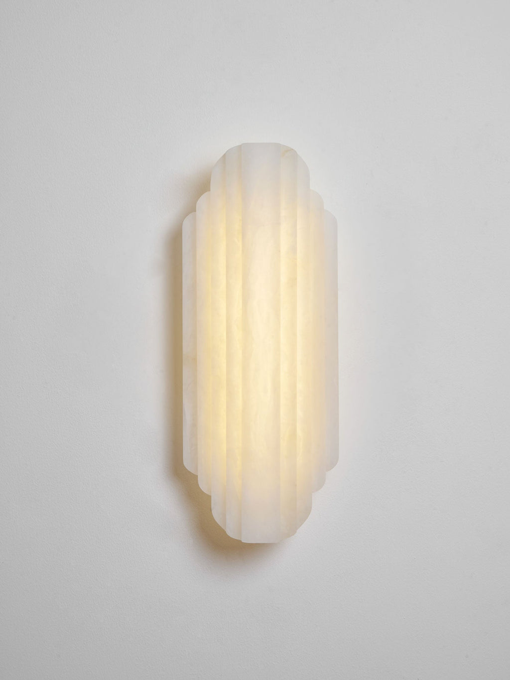 The shorter Clérachic alabaster sconce lit warm on a pale grey wall, its ribs glowing evenly from top to bottom.