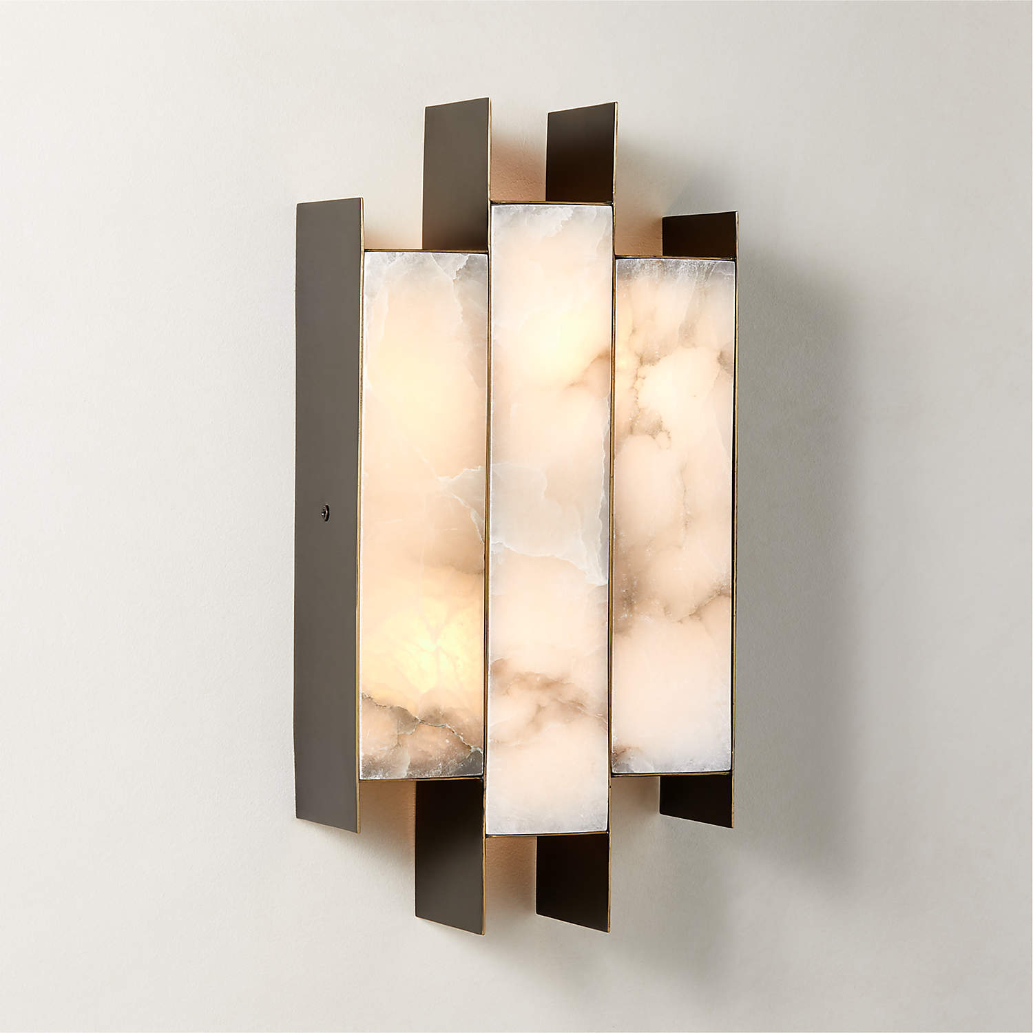 The Clérachic Alabaster Wall Sconce lit on a pale plaster wall, dark metal blades stepping past the glowing stone.