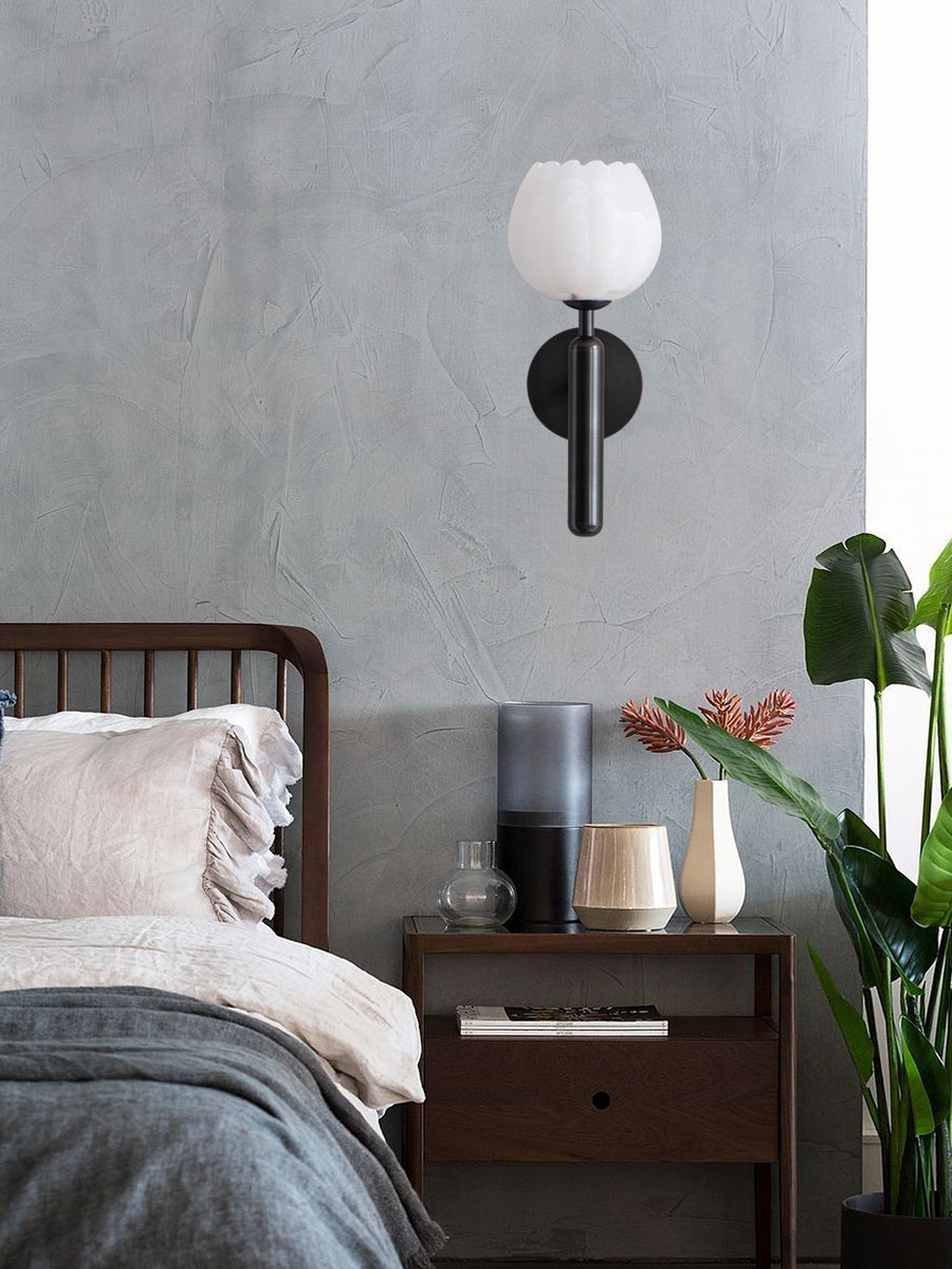 Black Clérachic alabaster wall sconce on a concrete-grey bedroom wall above a walnut nightstand and a tall green plant.
