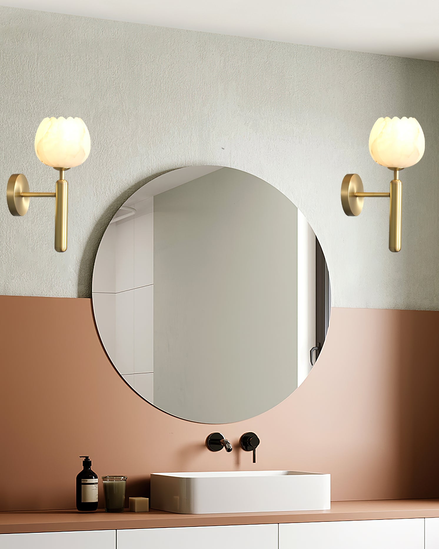 A pair of brass Clérachic alabaster wall sconces flanking a round mirror above a terracotta vanity, both tulip shades lit.