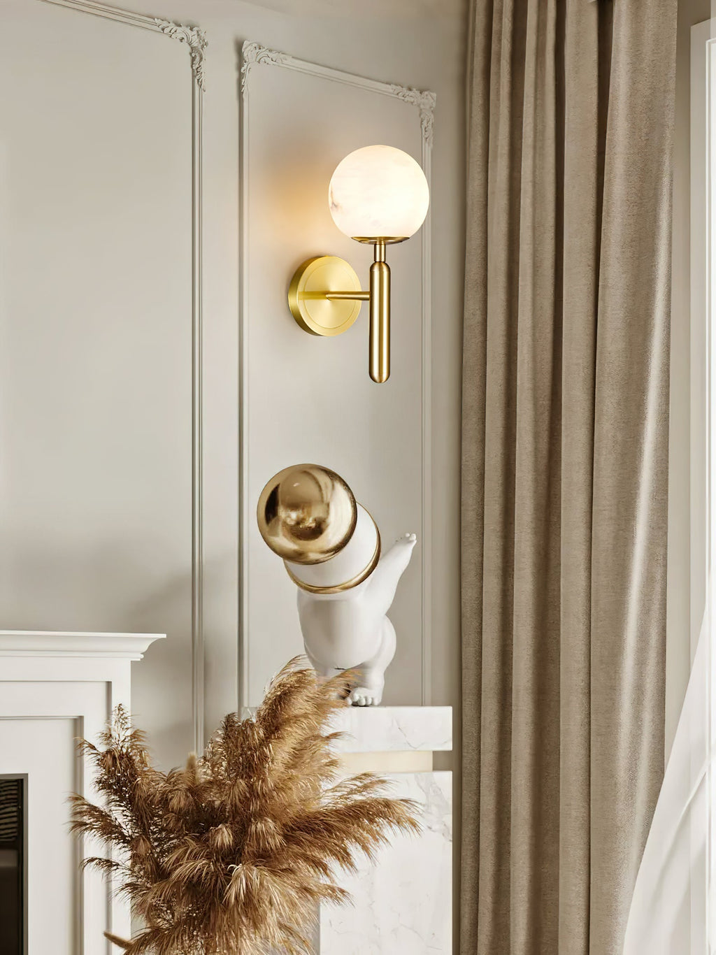 Clérachic alabaster wall sconce in brass, globe shade lit on a cream moulded wall above a mantel of dried pampas grass.