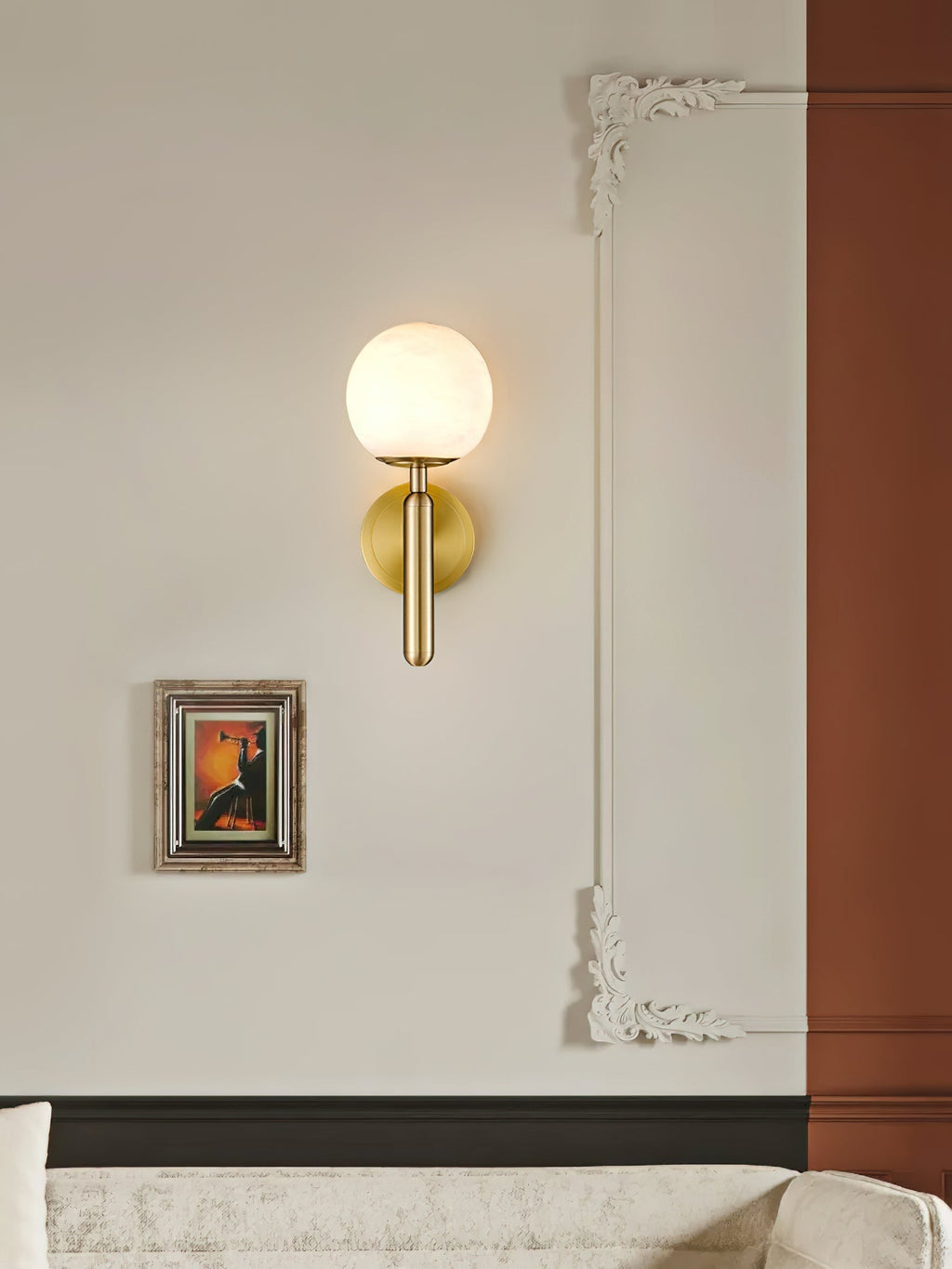 The Clérachic alabaster wall sconce in brass lights a cream drawing-room wall beside a small framed painting and moulding.