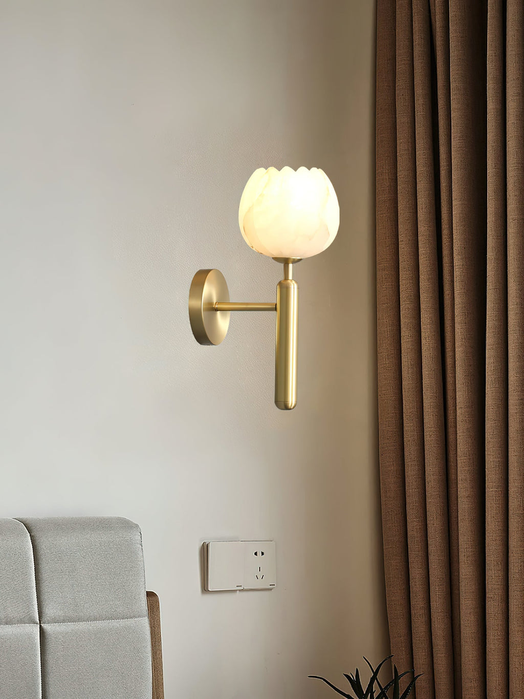 Brass Clérachic alabaster wall sconce lit beside a grey upholstered headboard, a brown linen curtain falling to its right.
