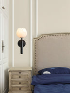 Black Clérachic alabaster wall sconce lit between a wardrobe and a nailhead headboard, tulip shade over a painted nightstand.