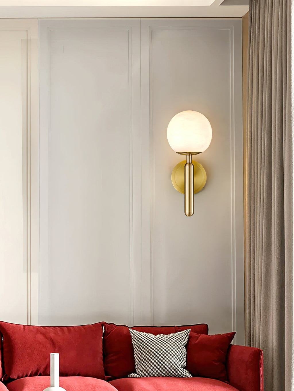 Brass Clérachic alabaster wall sconce glowing on a panelled wall above a deep red velvet sofa and a gingham cushion.