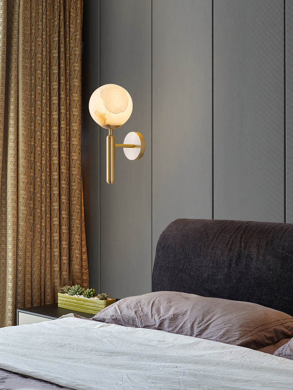 Brass Clérachic alabaster wall sconce lit on a grey panelled bedroom wall beside a patterned gold curtain and succulents.