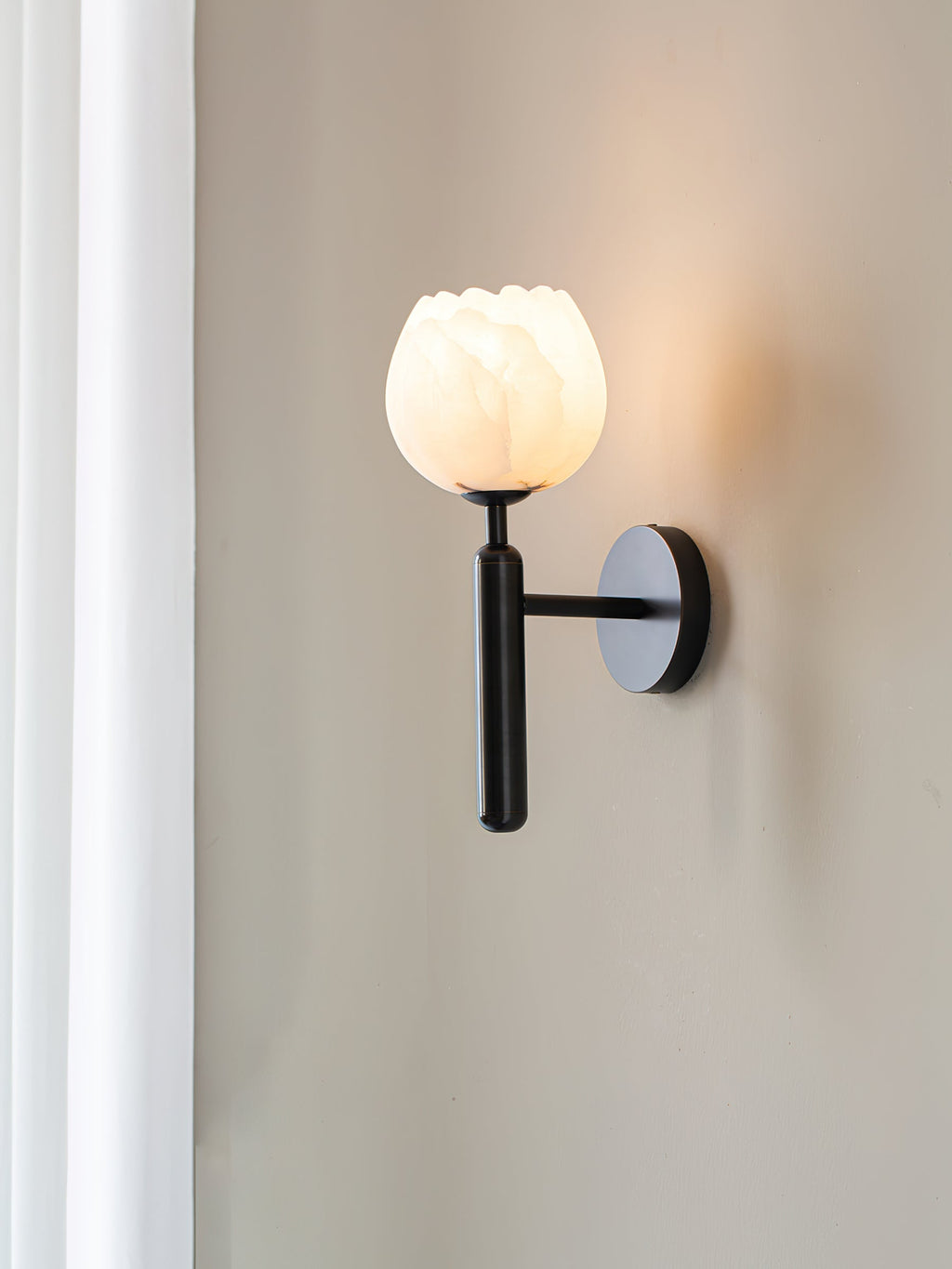 Clérachic alabaster wall sconce in black beside a white curtain, the arm holding the lit tulip shade clear of the wall.