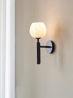 Clérachic alabaster wall sconce in black beside a white curtain, the arm holding the lit tulip shade clear of the wall.