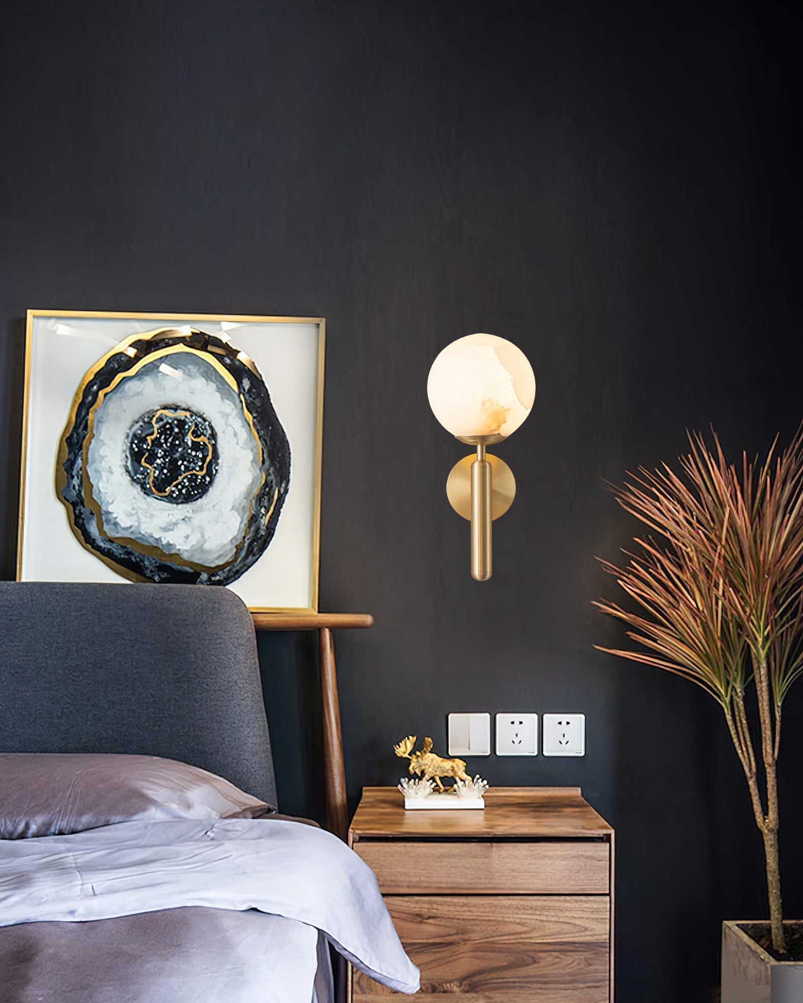 Brass Clérachic alabaster wall sconce on a near-black bedroom wall between framed agate art and a walnut nightstand.