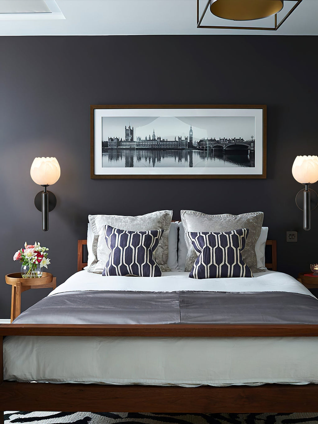 Two black Clérachic alabaster wall sconces lit either side of a framed London panorama above a walnut bed.