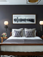 Two black Clérachic alabaster wall sconces lit either side of a framed London panorama above a walnut bed.