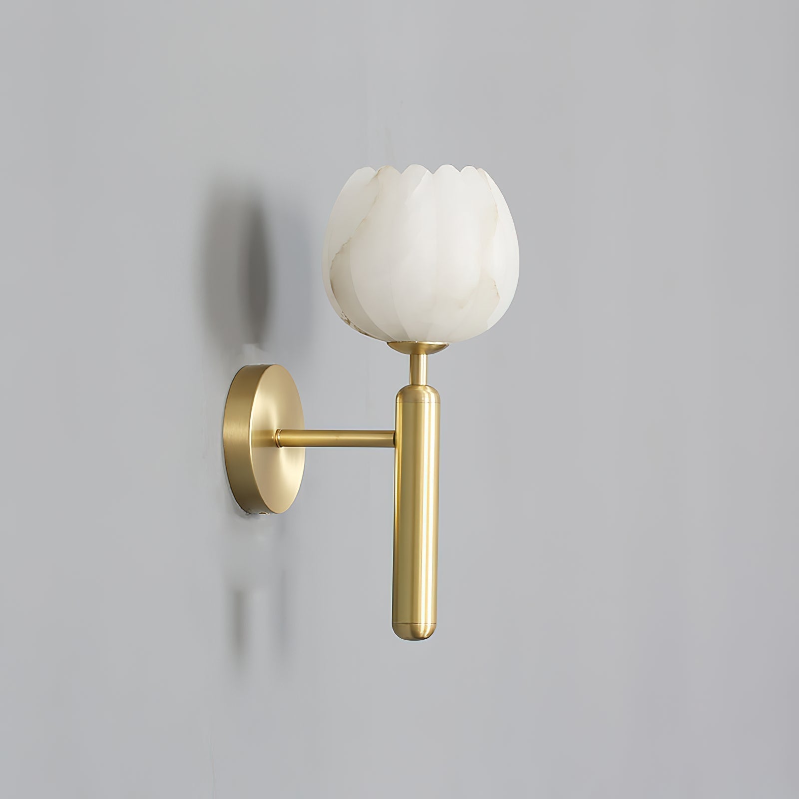 Angled view of the brass Clérachic alabaster wall sconce on a plain grey wall, the lit cup shade casting a soft shadow.