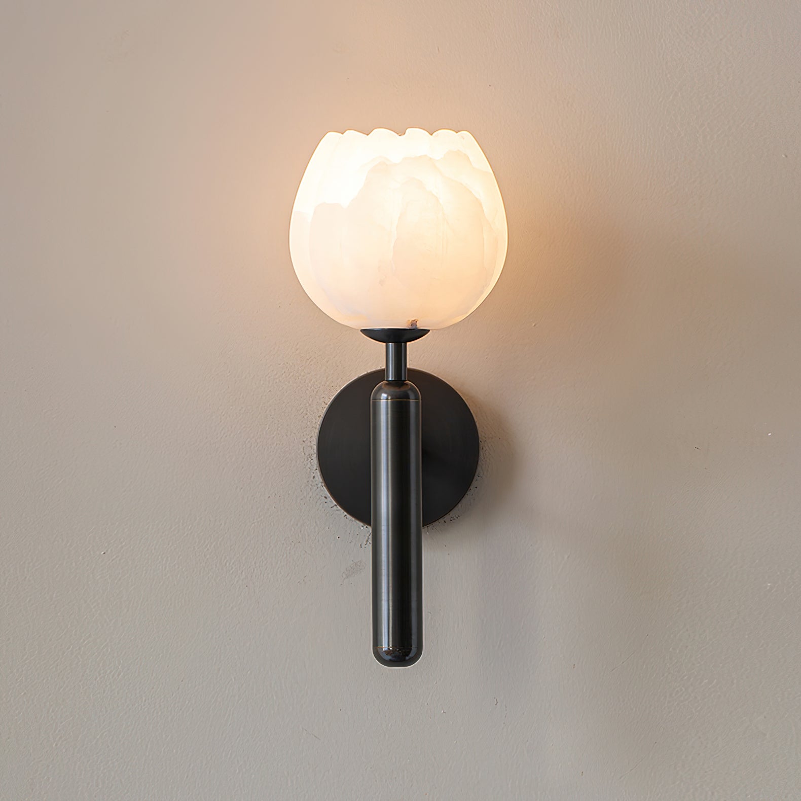 Black Clérachic alabaster wall sconce switched on, the stone cup warm gold and a halo of light spread across the wall.