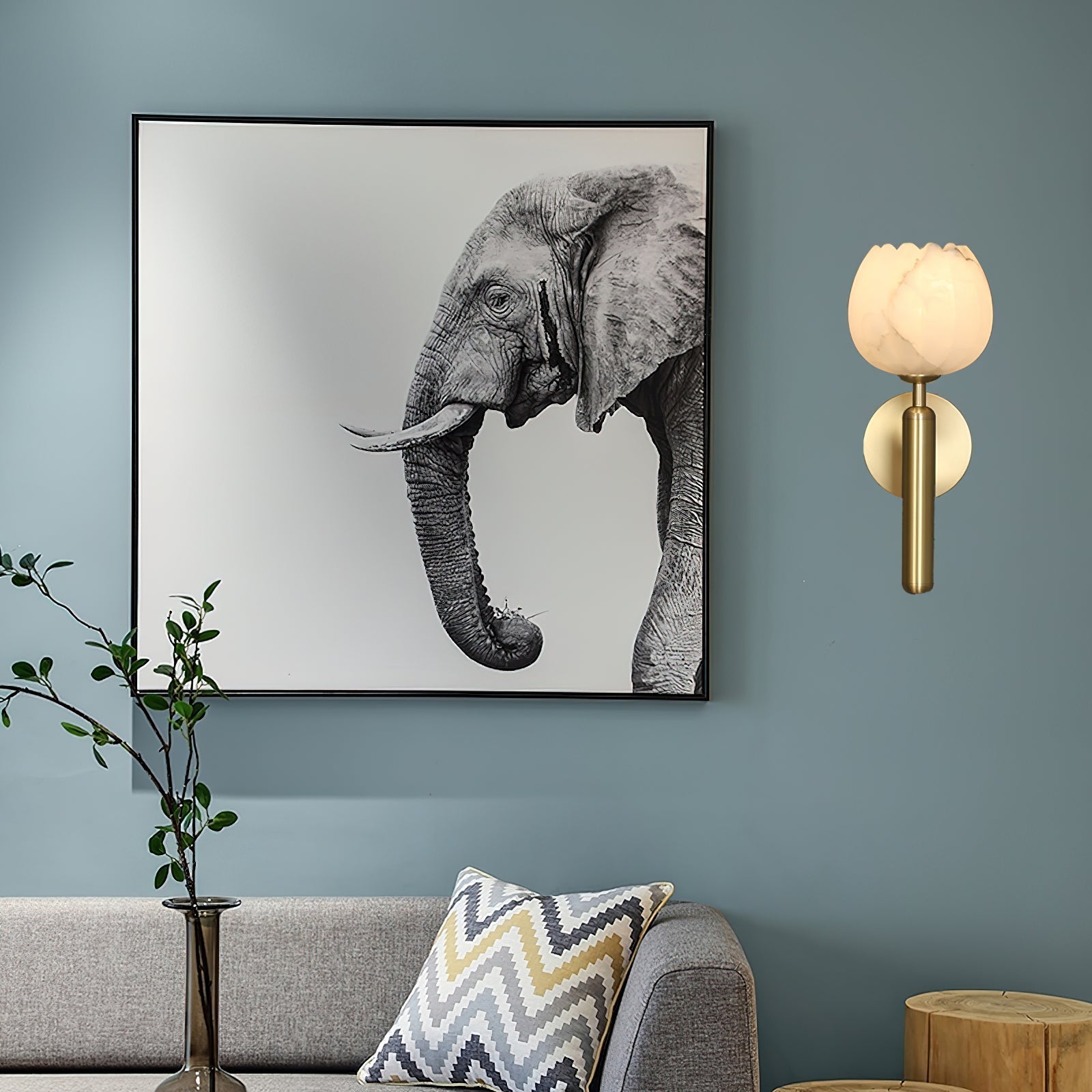 Brass Clérachic alabaster wall sconce lit on a blue-grey living-room wall beside a framed elephant print and grey sofa.