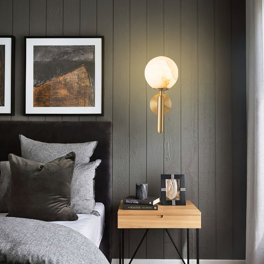 Brass Clérachic alabaster wall sconce lit on a dark timber-panelled bedroom wall above a nightstand stacked with books.