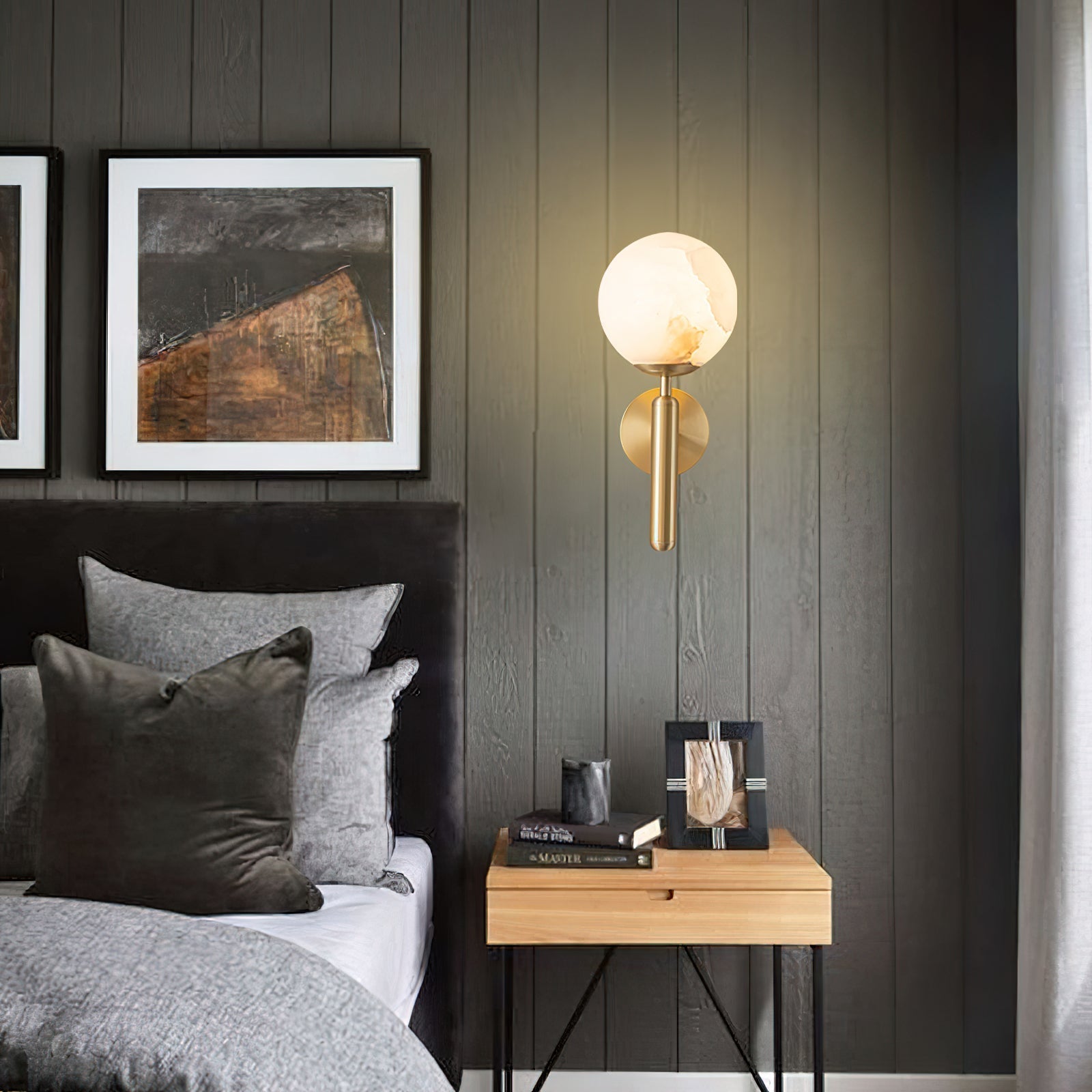 Brass Clérachic alabaster wall sconce lit on a dark timber-panelled bedroom wall above a nightstand stacked with books.