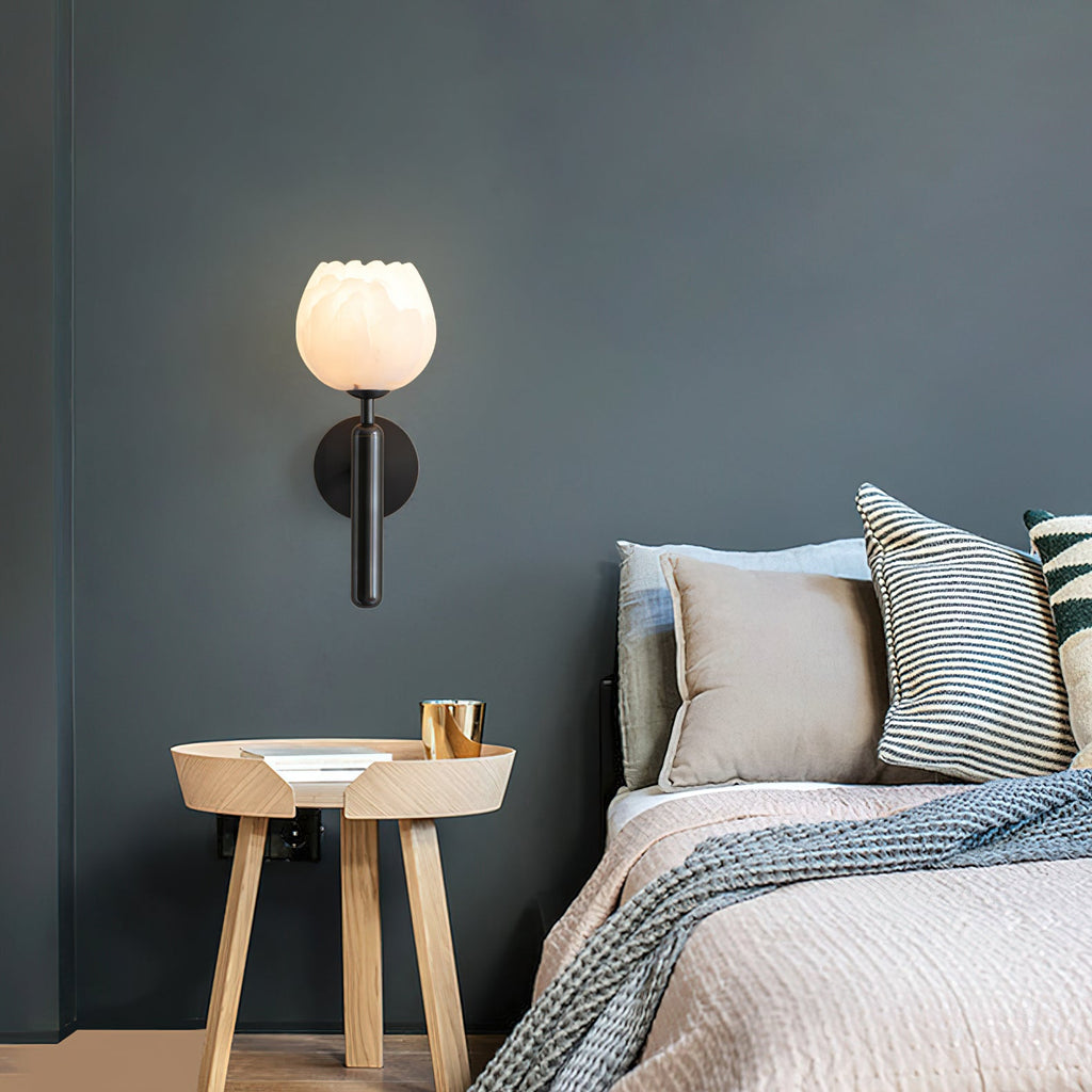 Black Clérachic alabaster wall sconce lit on a teal-grey bedroom wall above a round wooden side table beside the bed.