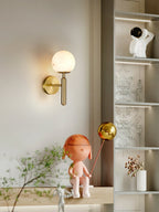 Brass Clérachic alabaster wall sconce lit on a cream wall beside lit display shelving and a pink figurine with a balloon.