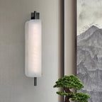 Clérachic Alabaster Wall Sconce lit on a pale linen wall beside a bonsai, the stone glowing warm with brass pins catching the light.