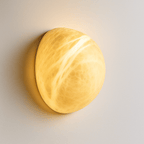 Clérachic pebble-shaped alabaster wall sconce lit warm, the veined stone glowing golden against a pale wall.