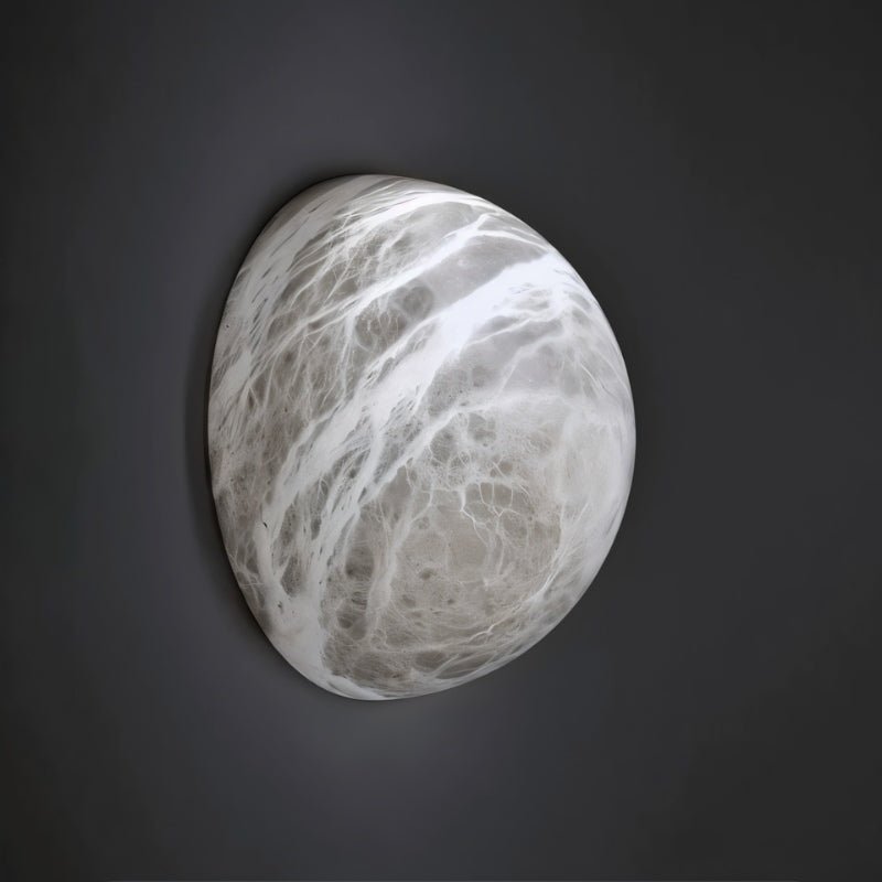 Clérachic pebble-shaped alabaster wall sconce with grey and white veining, mounted on a dark charcoal wall in the studio.