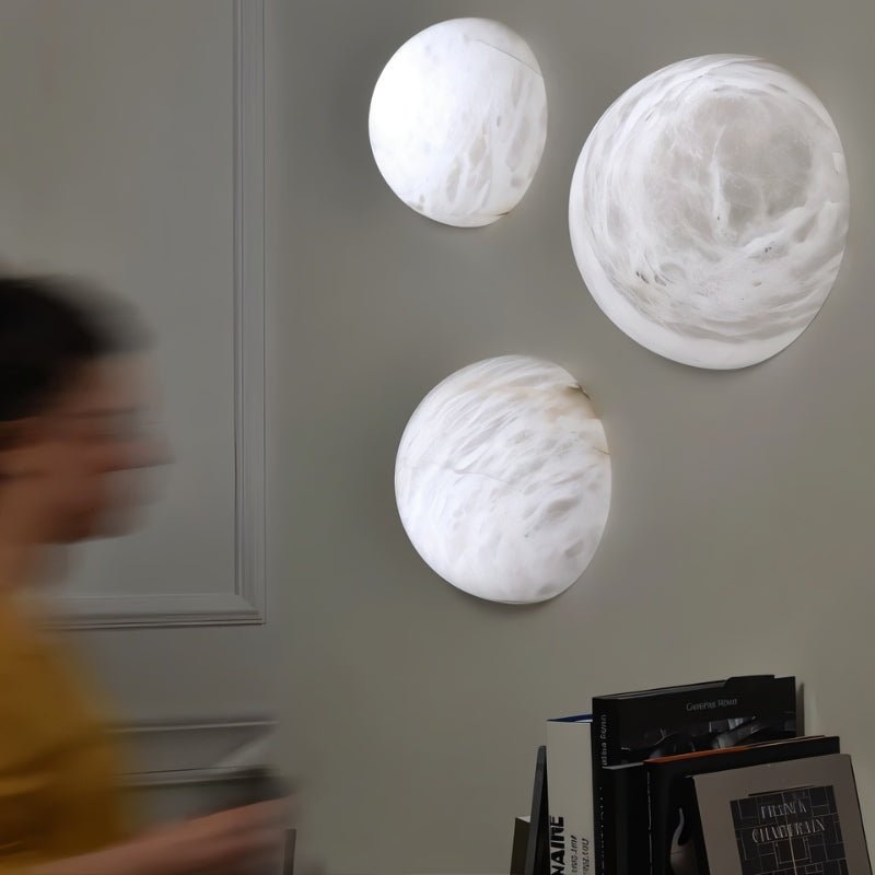 Clérachic three round white alabaster wall lights of different sizes on a grey panelled wall, above a stack of books in a room.