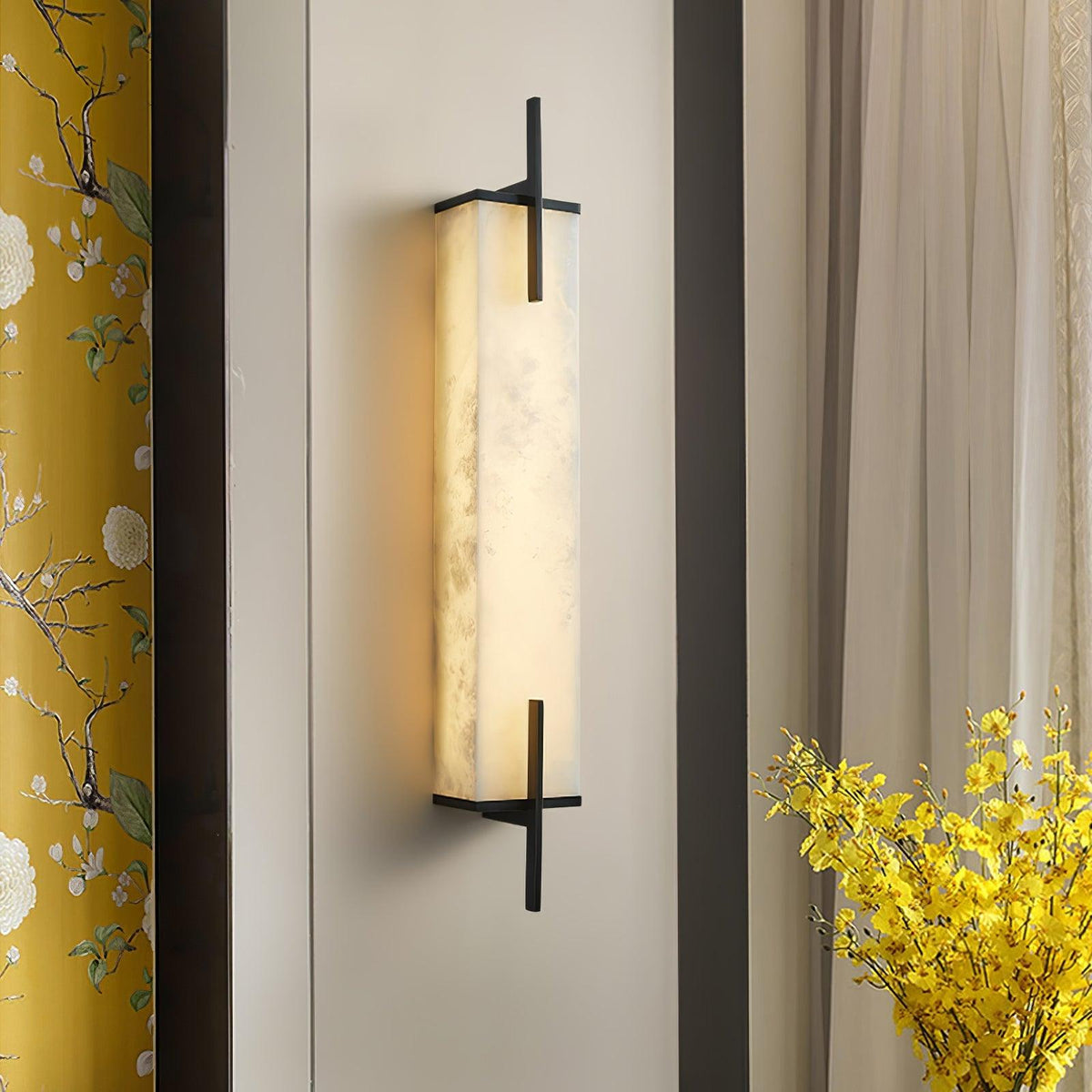 A lit Clérachic alabaster wall light with a black frame sits beside floral yellow curtains and a vase of yellow flowers.