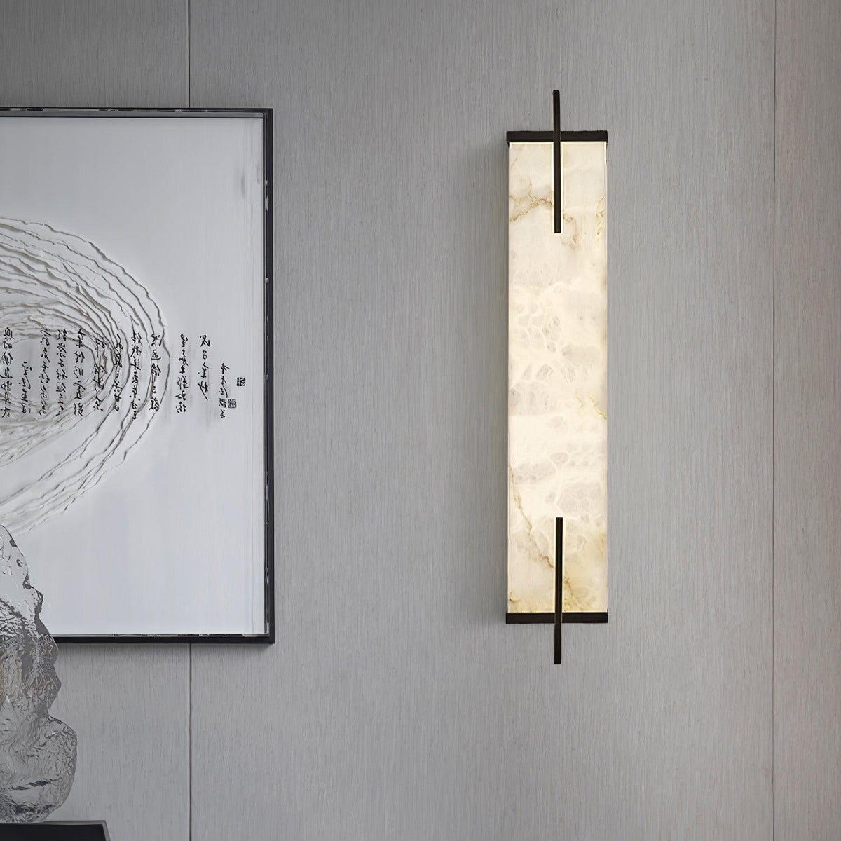 An unlit Clérachic alabaster wall light with black bars hangs on a grey panelled wall beside a framed calligraphy print.