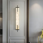 A Clérachic alabaster wall light with a black frame hangs inside a moulded panel on a pale wall, with yellow flowers at the edge.