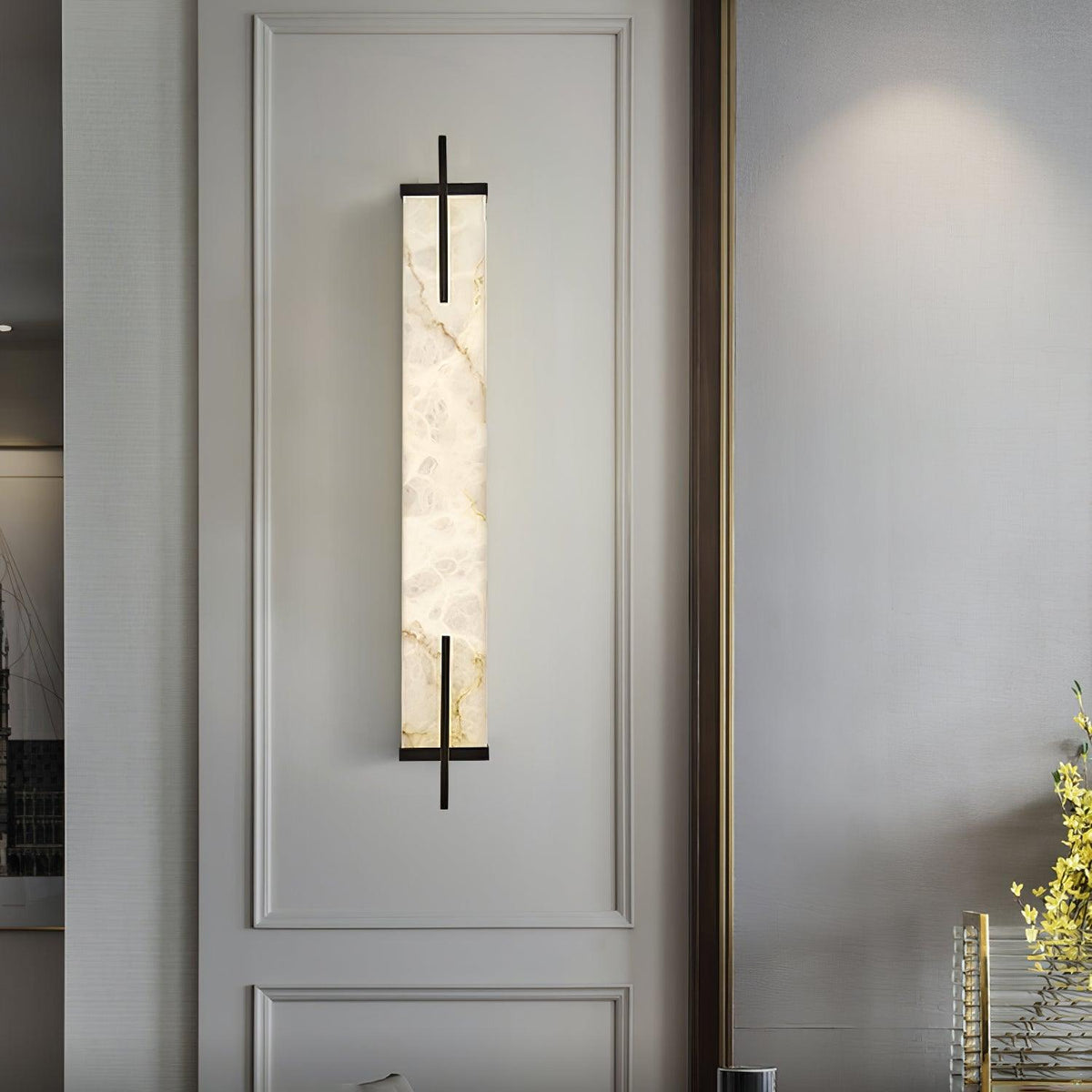 A Clérachic alabaster wall light with a black frame hangs inside a moulded panel on a pale wall, with yellow flowers at the edge.