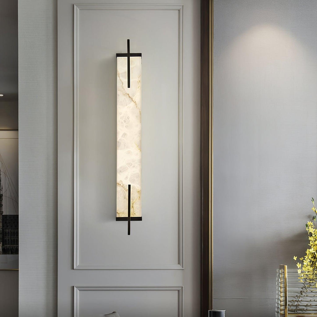 A tall Clérachic alabaster wall light hangs in a moulded wall panel, its veined stone glowing softly against pale grey.