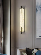 A Clérachic alabaster wall light glows beside a grey curtain and a pale abstract painting, with a sofa and cushions below.