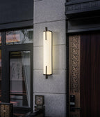Outside a dark entrance with a carved wooden door and glass panel, a lit Clérachic alabaster wall light glows against a stone-clad wall.
