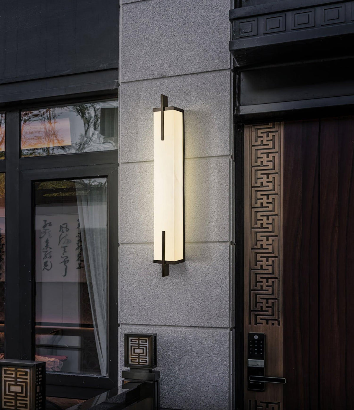 Outside a dark entrance with a carved wooden door and glass panel, a lit Clérachic alabaster wall light glows against a stone-clad wall.