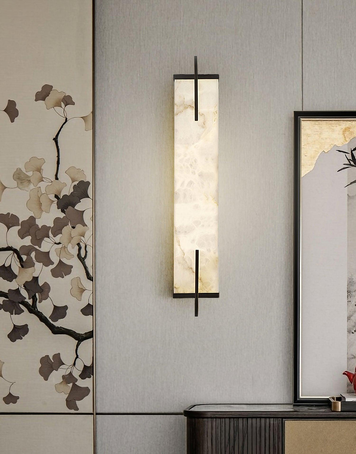 A Clérachic alabaster wall light hangs on a grey wall between a ginkgo-leaf wall panel and a framed landscape, above a dark console.