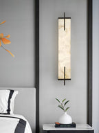 A Clérachic alabaster wall light with a black frame hangs above a bedside table with a white vase, a book and a corner of a grey headboard.