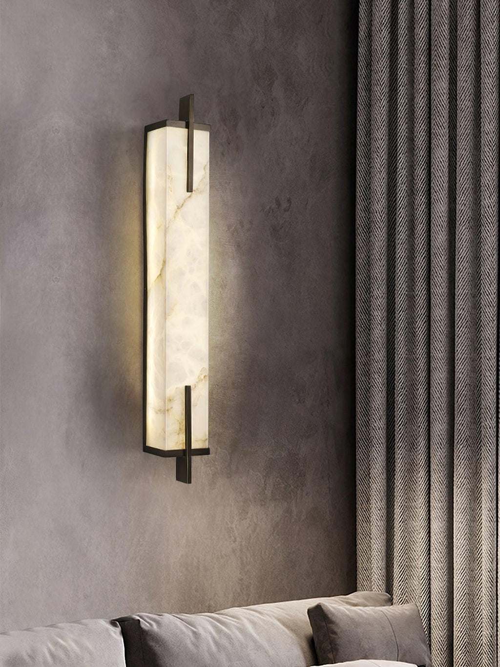 A softly lit Clérachic alabaster wall light on a grey wall beside heavy textured curtains, above the arm of a sofa.
