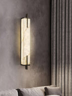 A softly lit Clérachic alabaster wall light on a grey wall beside heavy textured curtains, above the arm of a sofa.