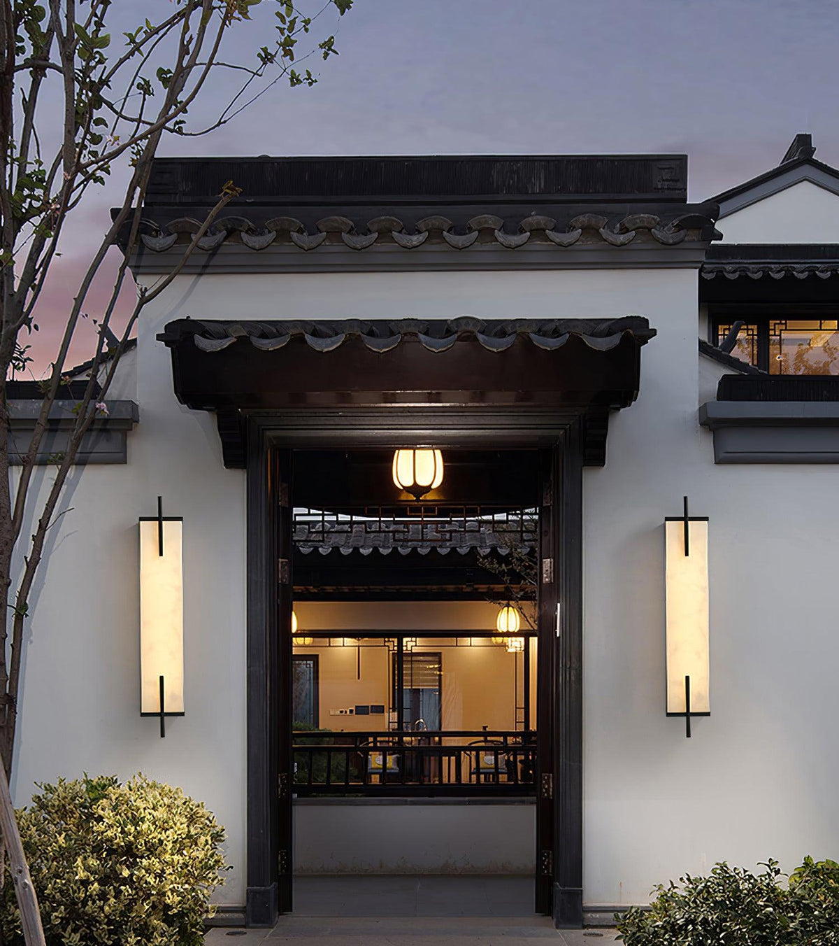 A pair of lit Clérachic alabaster wall lights flank a dark timber gateway on a white wall in a courtyard-style entrance at dusk.