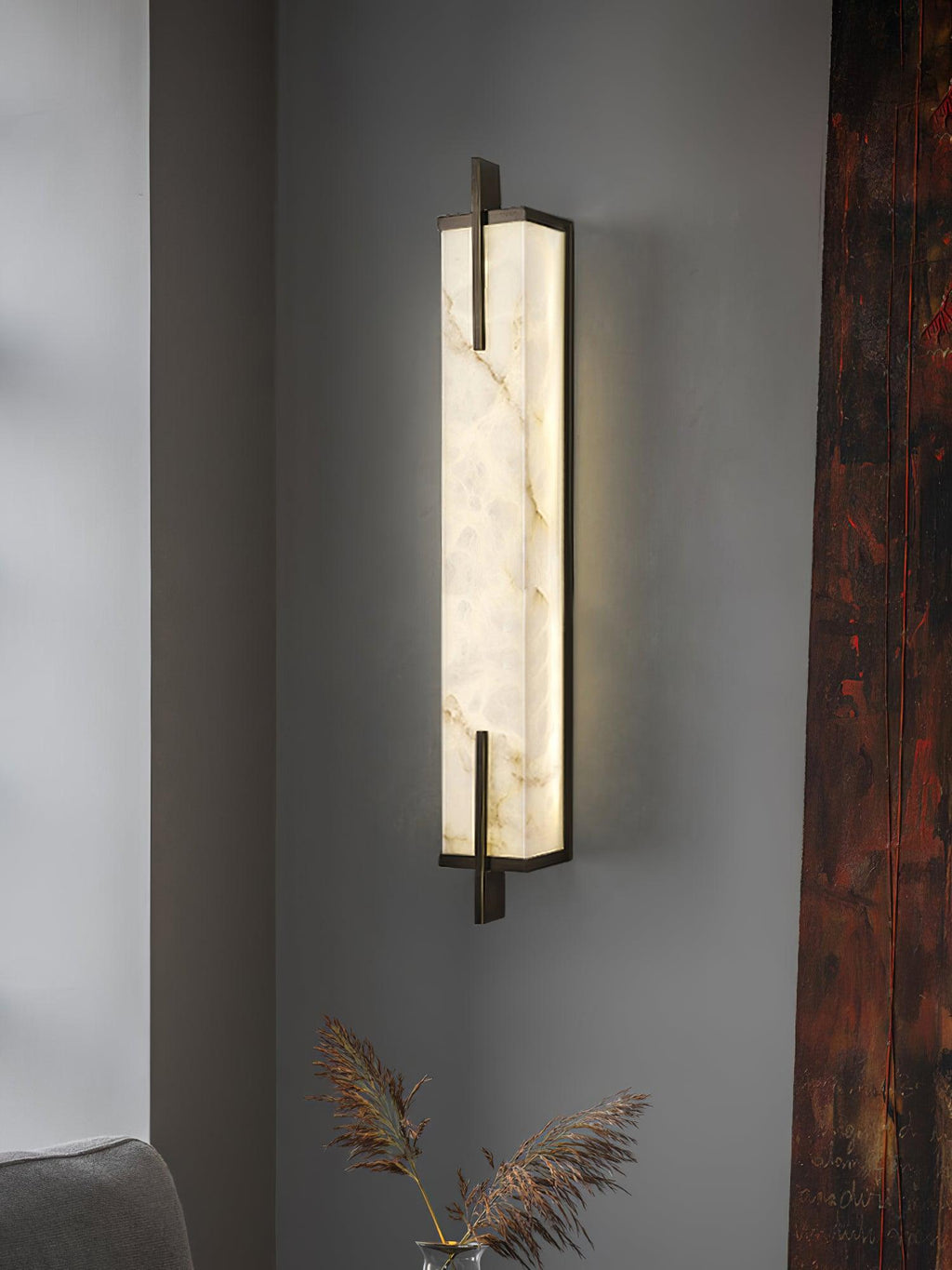 A Clérachic alabaster wall light glows on a grey wall beside a dark red timber post, with dried grasses in the corner.