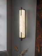 A Clérachic alabaster wall light glows on a grey wall beside a dark red timber post, with dried grasses in the corner.
