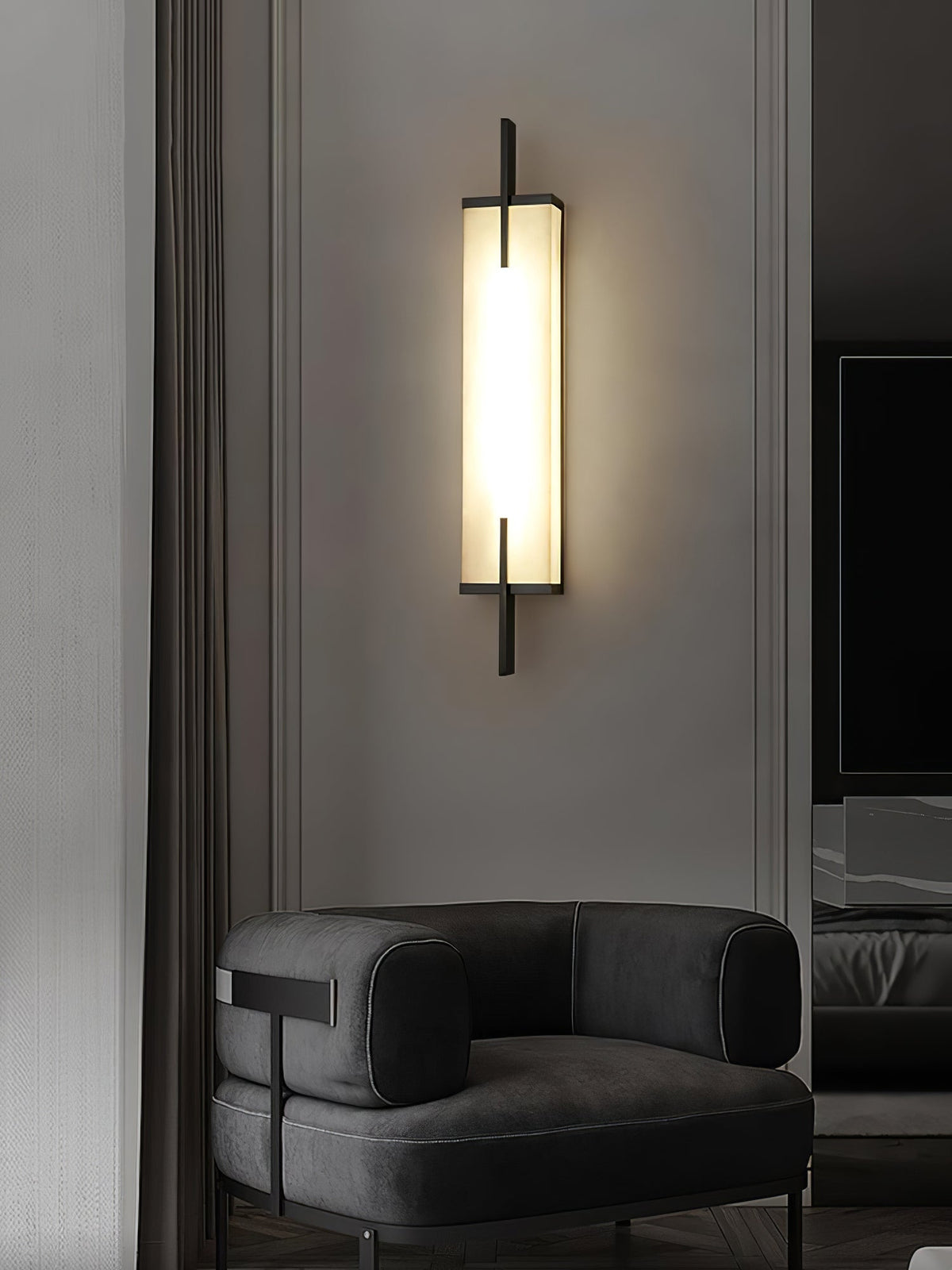 A lit Clérachic alabaster wall light with a black frame hangs on a pale wall above a dark grey armchair.