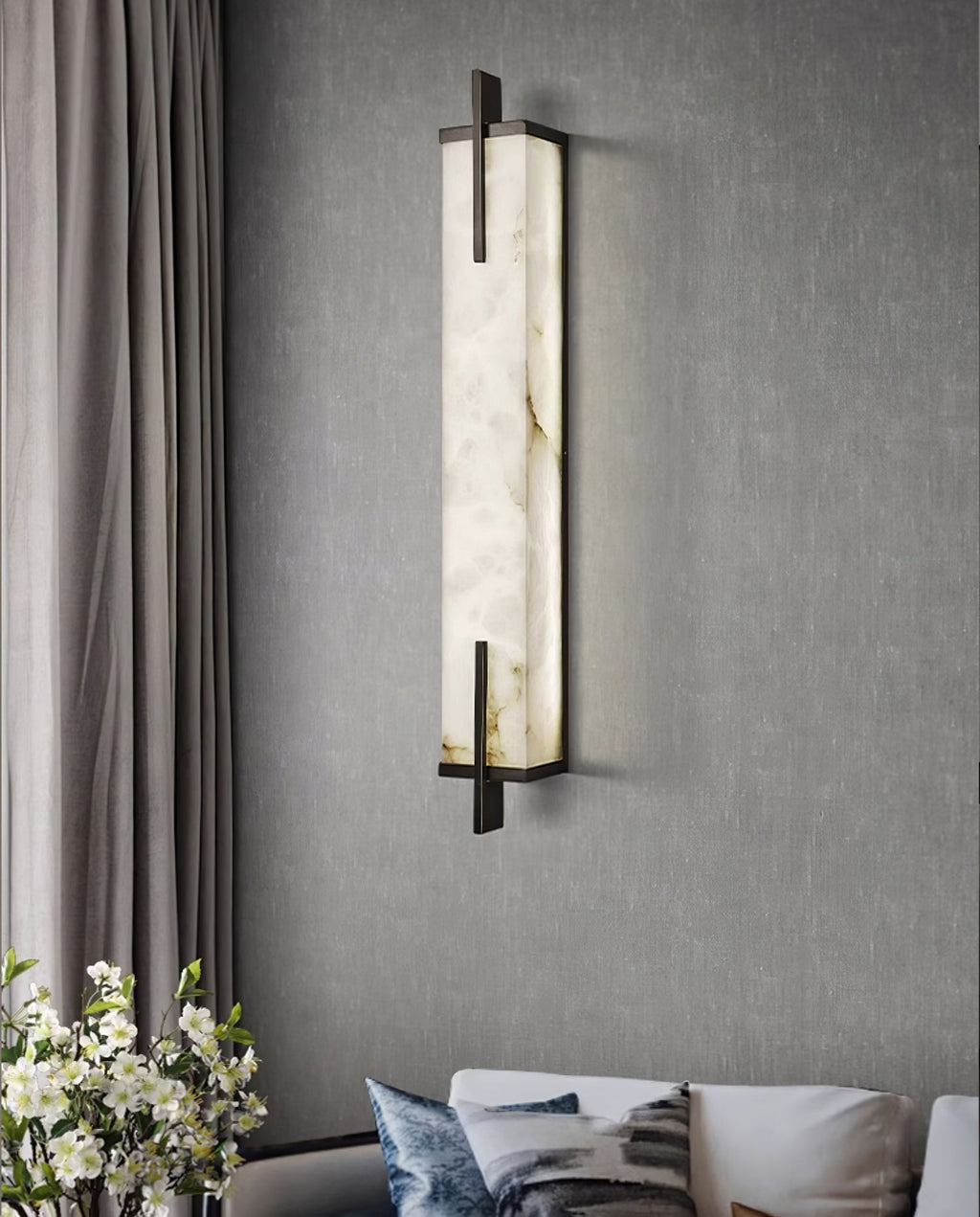 A Clérachic alabaster wall light with a black frame hangs on a grey wall beside textured curtains, with a bouquet of white flowers below.
