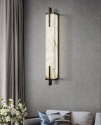 A Clérachic alabaster wall light with a black frame hangs on a grey wall beside textured curtains, with a bouquet of white flowers below.