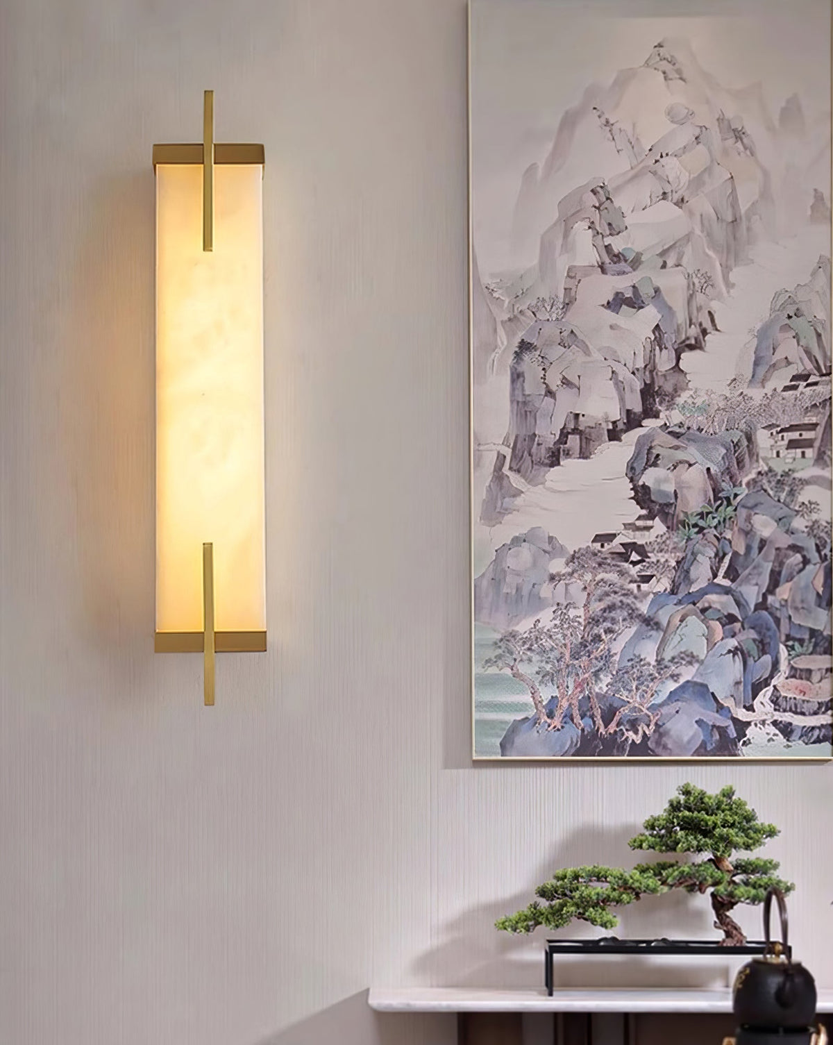 A lit Clérachic alabaster wall light with a gold-toned frame glows warmly beside a mountain landscape painting above a console.