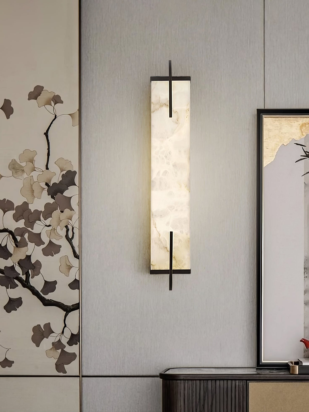 A Clérachic alabaster wall light with a black frame hangs against a ginkgo-leaf wall panel, beside a framed landscape and a dark console.