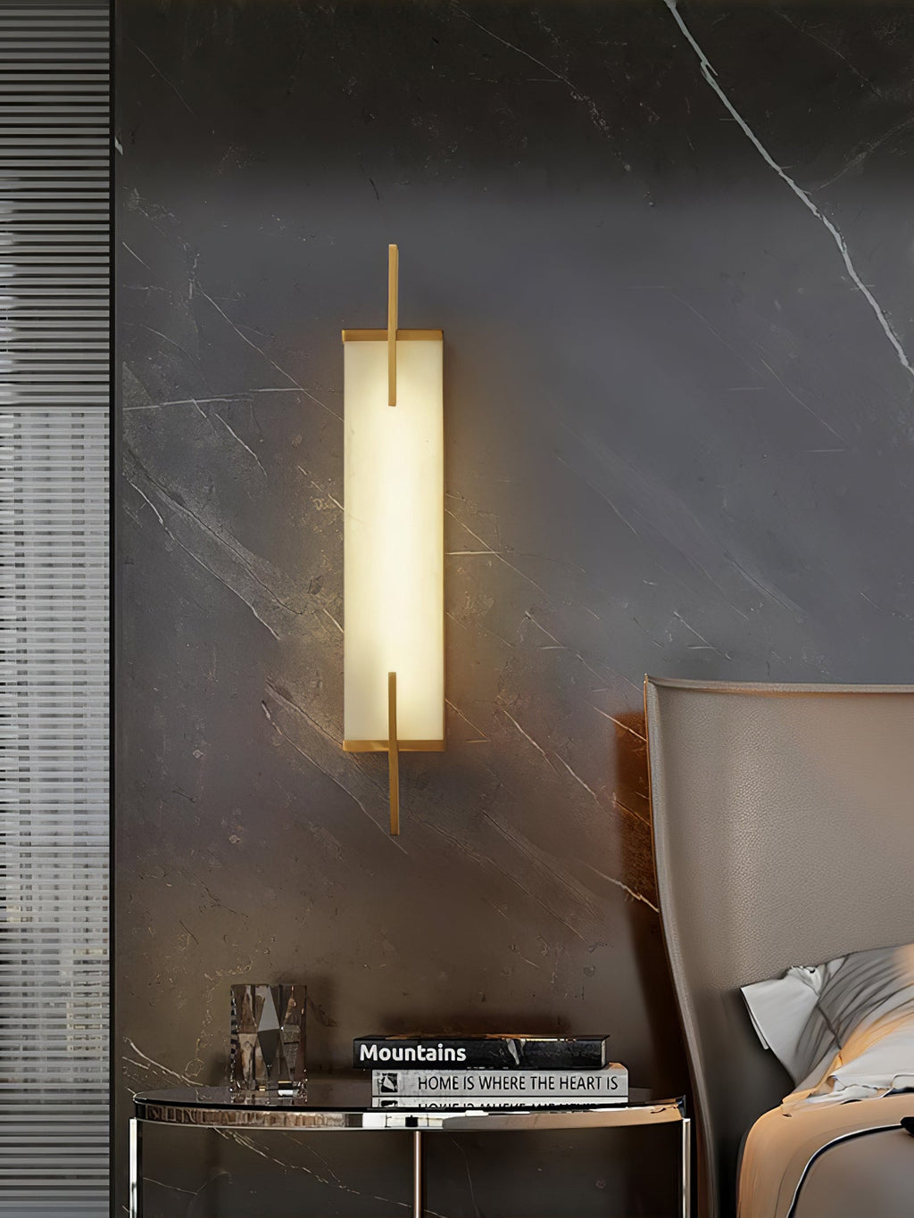 A lit Clérachic wall light with a gold-toned frame hangs on a dark marble-effect wall above a round side table and a grey chair.