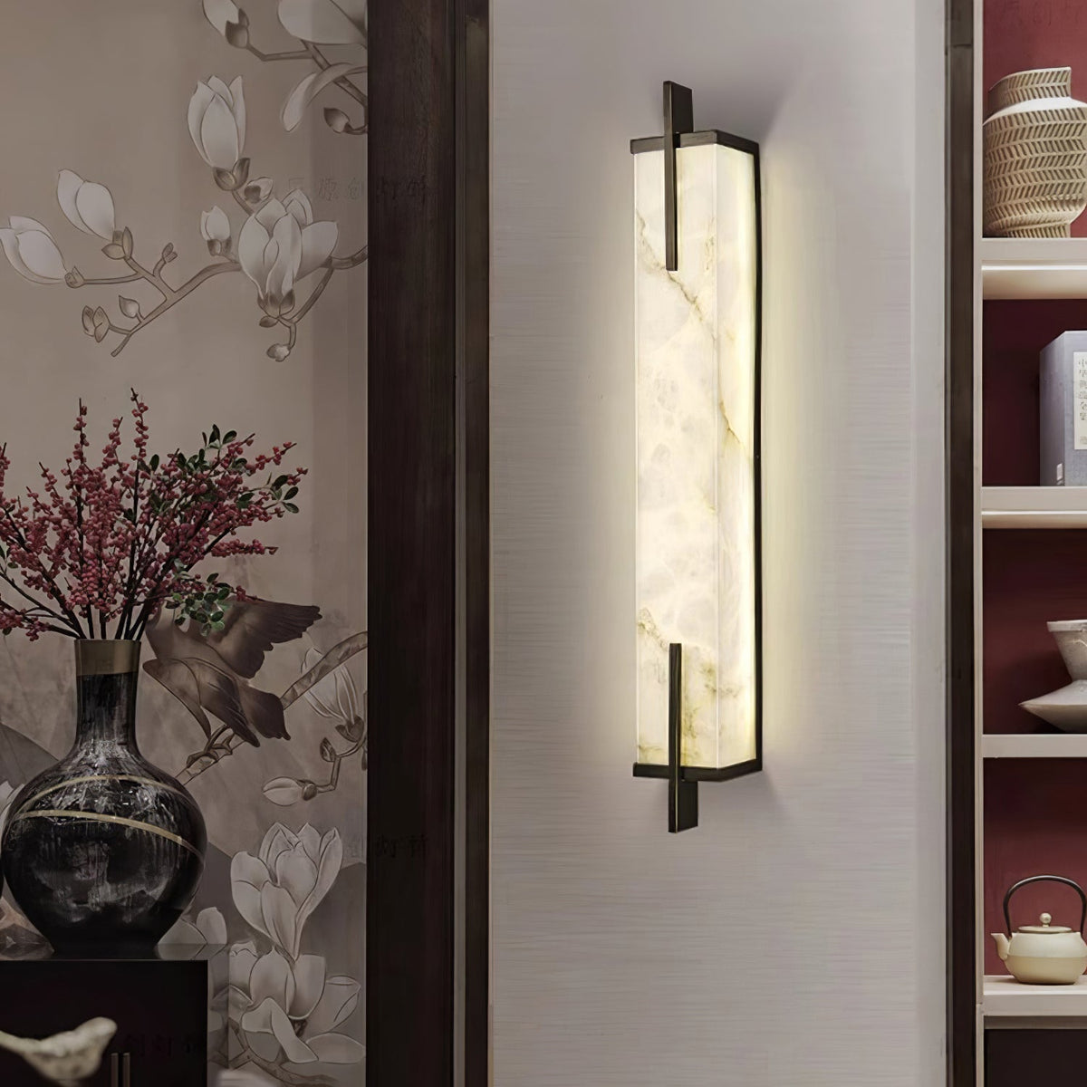 A Clérachic alabaster wall light glows on a pale wall beside a shelf unit, with a dark vase of berry branches and magnolia wallpaper.