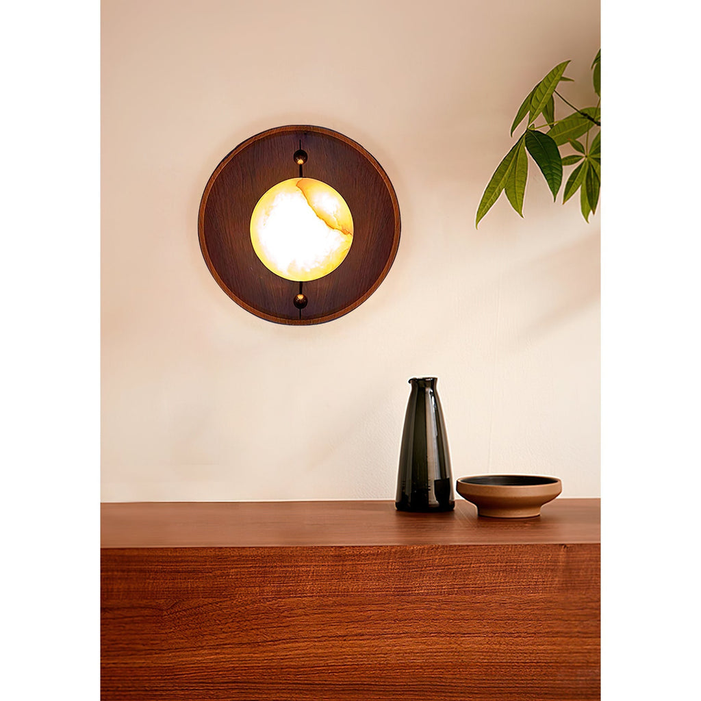 A Clérachic Alabaster Wall Sconce lit above a walnut sideboard, the stone reading like a small warm moon in its round wooden plate.