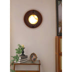 Round Clérachic Alabaster Wall Sconce lit in an entry corner, the walnut disc picked out against pale plaster beside a tall mirror.